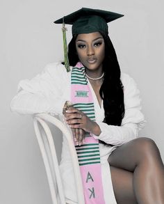 a woman in a graduation cap and gown sitting on a chair holding a pink sash