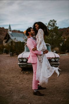 a man and woman standing next to each other in front of a pink car on dirt ground