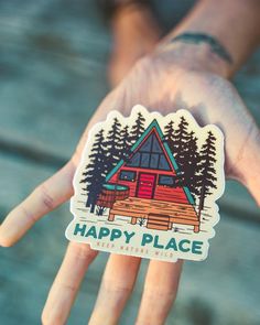 a hand holding a sticker that says happy place in front of a cabin with trees