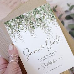 a person holding up a wedding card with greenery on the front and bottom, which reads save the date