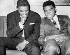 two men in suits sitting next to each other on a couch with their hands together