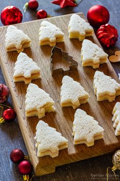 a wooden cutting board topped with cut up cookies next to christmas decorations and baubles