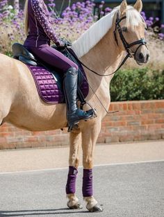 a woman riding on the back of a brown and white horse down a street next to purple flowers