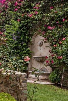 an outdoor fountain surrounded by pink flowers and greenery on the side of a stone wall