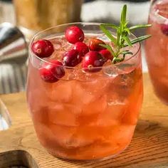 two glasses filled with ice and cranberries on top of a wooden table next to silverware