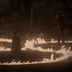 two people sitting on the ground in front of some fire and water with their backs to each other