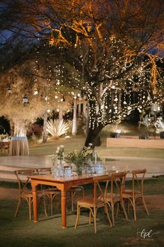 an outdoor dinner table set up with flowers and greenery on the table, surrounded by lights