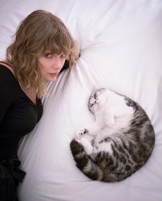 a woman laying in bed next to a cat on top of her stomach and looking at the camera