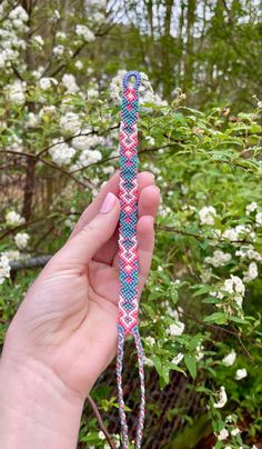 a hand holding up a pink and blue braided bracelet in front of white flowers