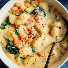 a white bowl filled with soup and vegetables on top of a blue cloth next to a spoon