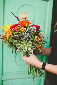 a woman holding a bouquet of flowers in front of a green door with an orange and yellow flower arrangement