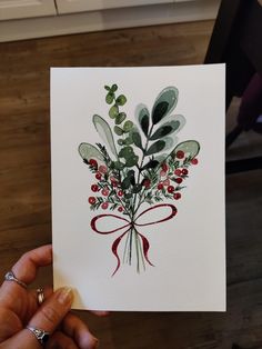 a person holding up a card with watercolor flowers and berries on it in front of a kitchen counter