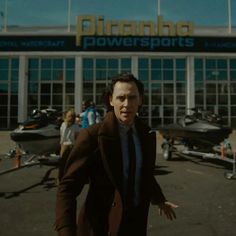 a man in a brown coat and tie walking towards a building with lots of boats