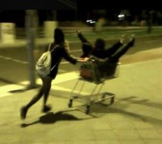 two people pushing a shopping cart down the street with their arms in the air and one person sitting on top of it