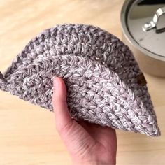 a hand is holding a crocheted hat on a table next to a clock