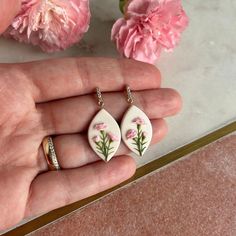 a person's hand holding two earrings with flowers painted on them and pink carnations in the background