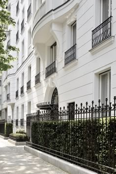 a white building with black iron fence and trees on the sidewalk in front of it