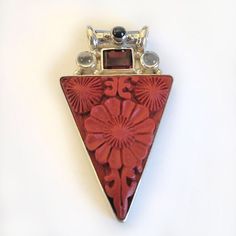 "Cinnabar, with a lovely floral design, has been bezel set in 925 Sterling Silver in this gorgeous bright red pendant.  Cinnabar is a resin that comes from the Laq tree in Asia. Once hardened, skilled artisans carve wonderful patterns in the medium.  This pendant also features a rectangle of Garnet, two rounds of Moonstone and a round of Black Star Diopside adorns the tube bale. The total length of this piece is 1 3/4\" long. Designed by Marianna and Richard and handmade by Balinese Artisans an Red Multi-stone Jewelry As Gift, Red Sterling Silver Jewelry With Inlay, Red Carved Jewelry Gift, Red Multi-stone Jewelry Gift, Red Carved Jewelry For Gifts, Unique Red Cabochon Jewelry, Red Multi-stone Jewelry For Gifts, Luxury Red Rectangular Pendant Jewelry, Spiritual Red Cabochon Jewelry