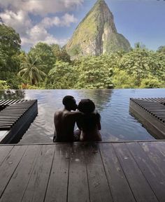 two people are sitting on the edge of a pool looking at mountains and trees in the distance