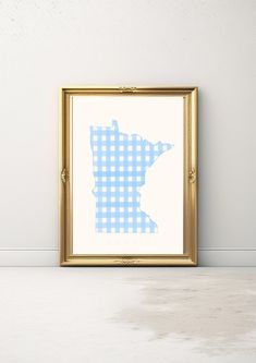 a blue and white gingham pattern in a gold frame on the floor next to a wall