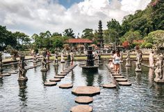 people are standing on stepping stones in the water at a park with fountains and statues