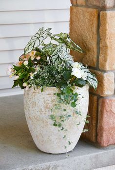 a planter with flowers and greenery in front of a brick wall on a porch