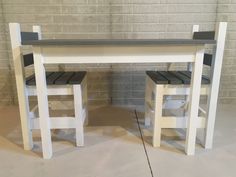 two wooden stools sitting next to a white table and bench on concrete floor in front of brick wall