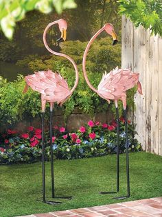 two pink flamingos are standing in the grass next to a fence and flower garden