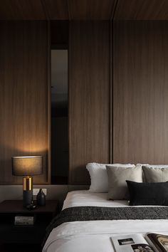 a bed with white sheets and pillows next to a lamp on a night stand in front of a wooden paneled wall