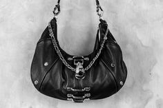 An ode to the timeless partnership of leather and chains, this leveled up black hobo bag is made from soft, premium genuine black leather and features a top shelf arrangement of silver hardware throughout.A centered, weighty metal industrial clip takes center stage, accompanied by an array of high contrast chromed out nailed rivets, centered feed through bars, and a draped top chain that doubles as a shoulder strap.Complete with a removable/adjustable leather and chain strap that can easily convert this purse from a shoulder bag to a comfortable crossbody bag, this design is an ode to the tough and timeless partnership of leather and chains.Bag is approx 13.5" x 7"Removable strap can extend up to 40" Leather Hobo Bag With Chain Strap, Edgy Leather Shoulder Bag With Metal Hardware, Silver Leather Shoulder Bag With Hardware, Black Edgy Bags With Palladium Hardware, Edgy Black Bags With Palladium Hardware, Edgy Black Shoulder Bag With Palladium Hardware, Punk Leather Shoulder Bag With Gunmetal Hardware, Gothic Leather Shoulder Bag With Metal Hardware, Gothic Leather Shoulder Bag With Hardware