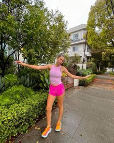 a woman in pink shirt and shorts standing on sidewalk next to bushes with her arms out