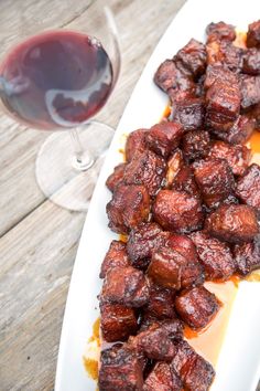 a white plate topped with meat next to a glass of red wine on top of a wooden table