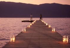 two people standing on the end of a dock with candles lit up in front of them