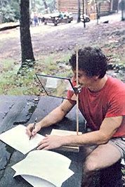 a man sitting at a picnic table writing on a piece of paper while holding a pen