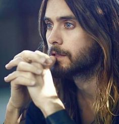 a man with long hair and blue eyes is holding his hand to his chin while looking at the camera