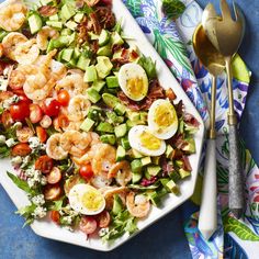 a salad with shrimp, eggs and tomatoes on a white platter next to two spoons