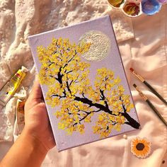 a person holding up a painting with yellow flowers and paintbrushes next to it