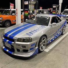 a silver car with blue stripes parked in a parking lot next to other cars and gas pumps