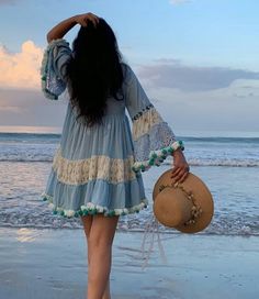 a woman in a blue dress is walking on the beach with her hat over her head