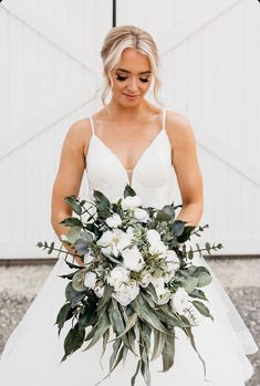 a bride holding her bouquet in front of a barn door with white flowers and greenery