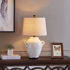 a white vase sitting on top of a wooden table next to a lamp and framed pictures