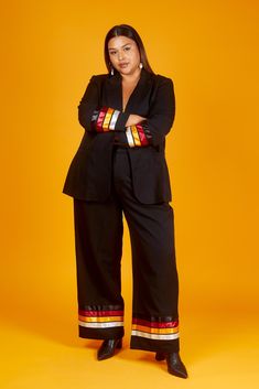 a woman standing with her arms crossed in front of an orange background wearing black pants and a blazer