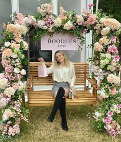 a woman sitting on a wooden bench in front of a floral arch holding a shopping bag