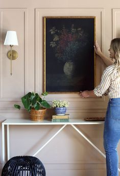 a woman standing in front of a painting on a wall next to a table with potted plants