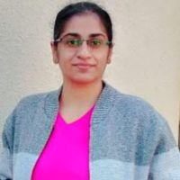 a woman with glasses standing in front of a wall wearing a pink shirt and blue cardigan