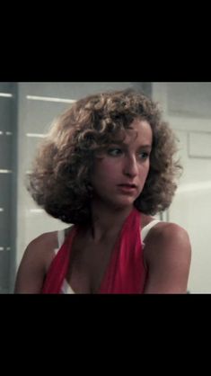 a woman with curly hair in a red dress looking at the camera while standing next to a mirror