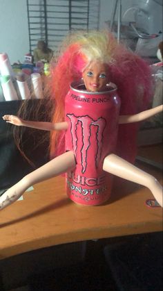 a doll with blonde hair is posed in a pink monster energy drink can as she sits on a desk