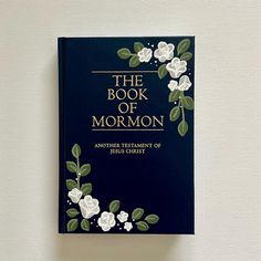 the book of mormon with white flowers and green leaves on blue cover against a white wall