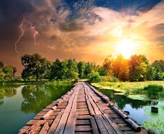 a wooden bridge that is over water with lightning in the sky and trees behind it