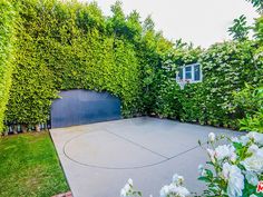 an outdoor basketball court surrounded by bushes and flowers in front of a house with a blue gate